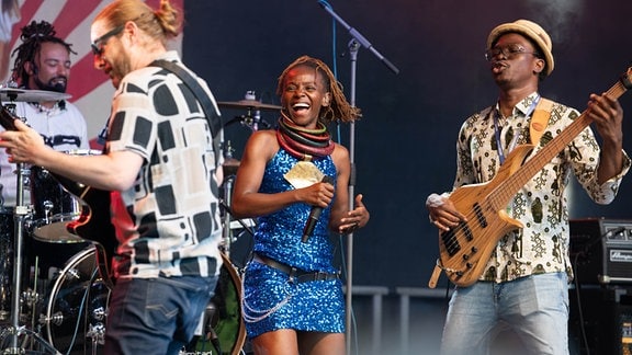 Musicians on a stage at the Rudolstadt Festival, two guitarists with sunglasses, a drummer and a beaming woman with chokers and a short blue glitter dress.